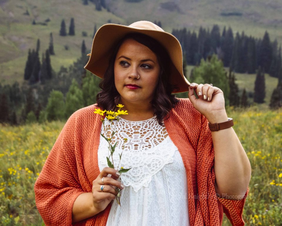 Portrait of a woman holding a yellow flower in front of a tree-lined landscape. She is wearing a floppy hat, white shirt, and coral colored sweater.